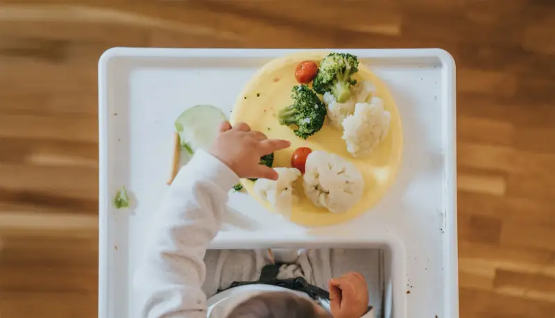 Zdrowe drugie śniadanie dla roczniaka: brokuły, kalafior i pomidorki na żółtym talerzyku. Dziecko sięga po jedzenie.