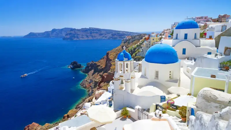 Santorini wycieczki: białe domy z niebieskimi kopułami na klifie nad błękitnym morzem. Statek płynie w oddali.