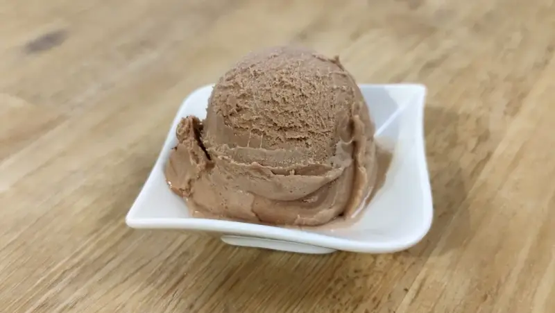 Ein Löffel cremiges Nougat Eis in einer weißen Schale auf einem Holztisch.