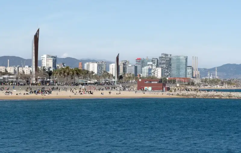 Ludzie odpoczywają na plaży, a w tle widać nowoczesne budynki Barcelony. To miasto leży nad Morzem Śródziemnym.
