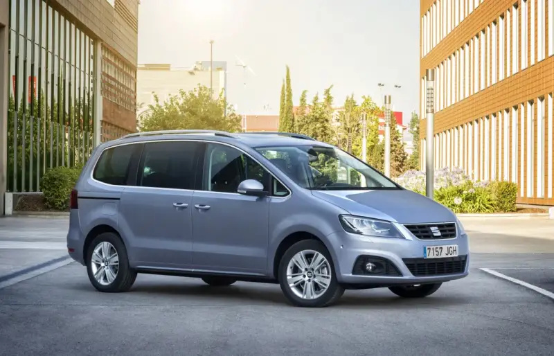 Srebrny Seat Alhambra, rodzinny van, parkuje przed nowoczesnym budynkiem z brązową elewacją.