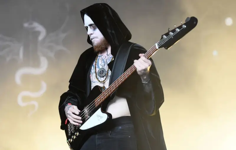 Le bassiste d'un groupe metal connu, maquillé et tatoué, joue sur scène, drapé dans une cape noire.