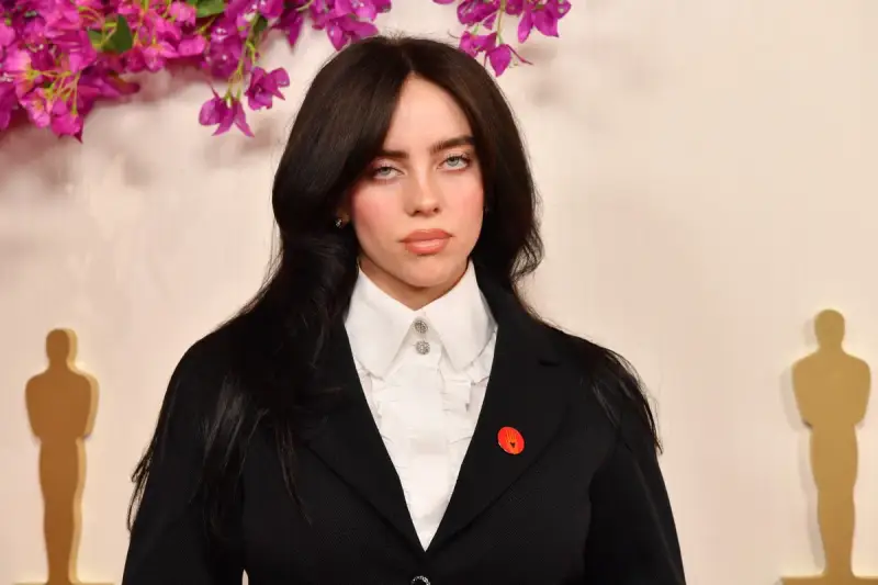 Billie Eilish z charakterystycznymi sanpaku eyes, w czarnym żakiecie i białej koszuli, na tle ozdobnych kwiatów i złotych statuetek.