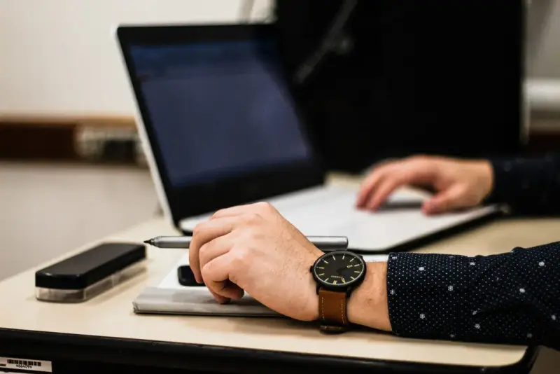 Dłoń z zegarkiem trzyma długopis nad dokumentem, obok laptop. Czas na skargę na przewlekłość postępowania opłatę.