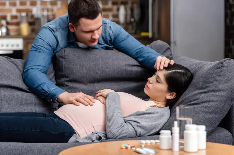 Zmartwiony partner troskliwie dotyka czoła ciężarnej kobiety, która leży na kanapie. Na stoliku leki, sugerujące problemy zdrowotne, być może angina w ciąży.