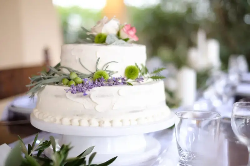 Dwupoziomowy tort weselny ozdobiony kwiatami, ziołami i oliwkami. Delikatne kremowe wykończenie i naturalne dekoracje podkreślają wyjątkowe smaki tortów weselnych.
