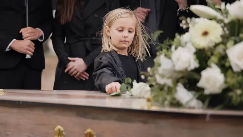 Smutne dziecko na pogrzebie, dotyka kwiatów na trumnie. W tle dorośli w czerni. Czy to dziecko wierzy w przesądy?