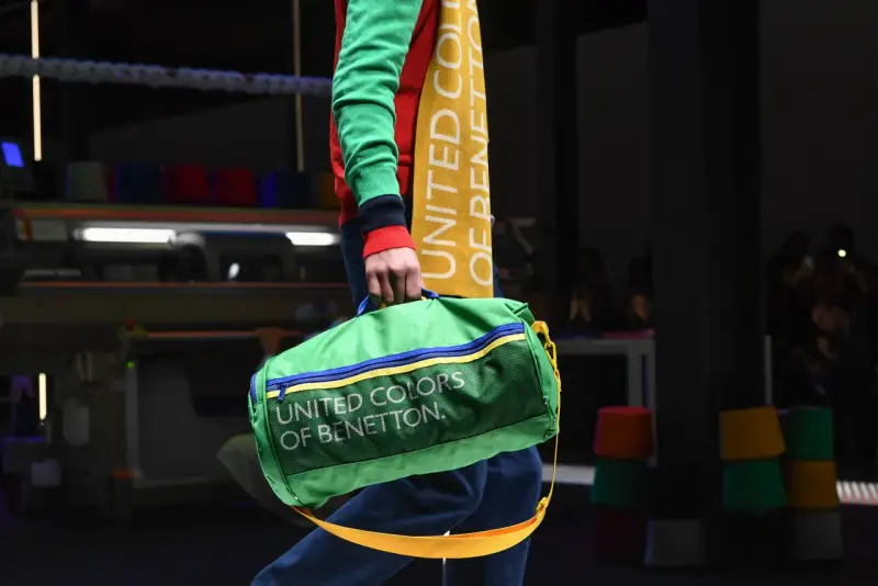 Zielony worek z napisem "UNITED COLORS OF BENETTON" niesiony przez modela w kolorowym swetrze.