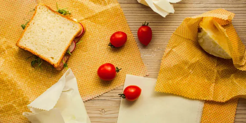 Un sandwich, des tomates et un morceau de fromage sont présentés sur des bee wraps maison à pois rouges sur fond de bois.