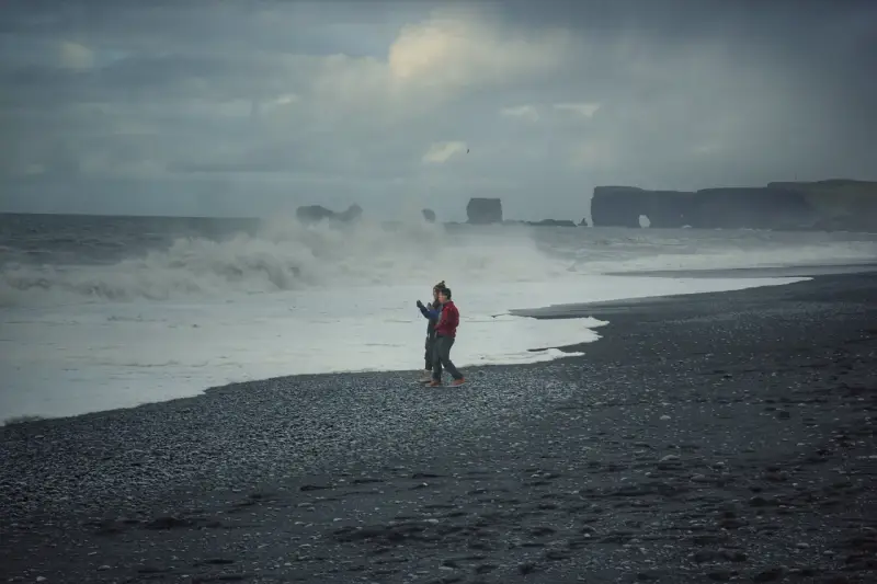 Para spaceruje po czarnej plaży Islandii, podczas gdy fale rozbijają się o brzeg.