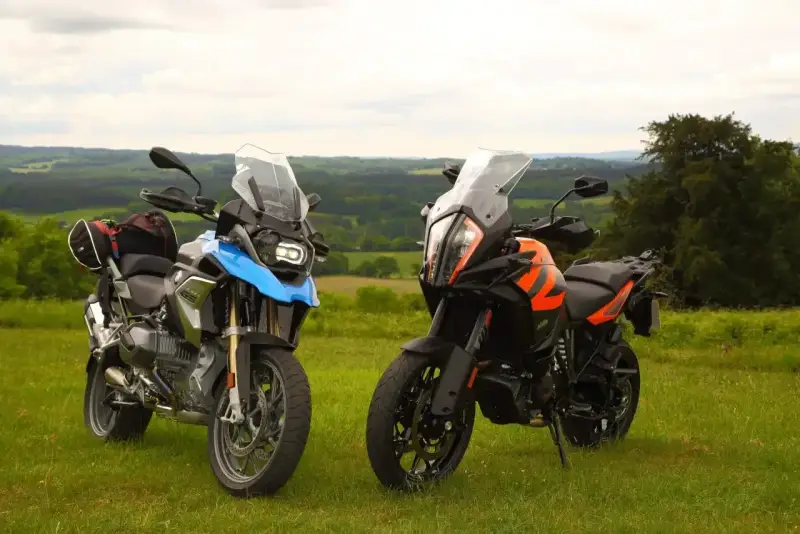 Dwa motocykle turystyczne, w tym pomarańczowy KTM 1290 Super Adventure, stoją na trawie z widokiem na zielone wzgórza.