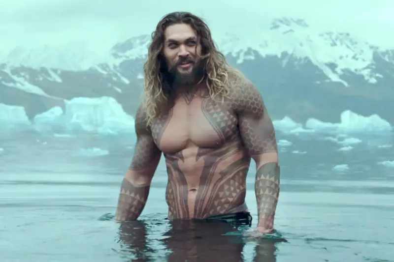 Jason Momoa w wodzie, z tatuażami, jak w filmach i serialach. W tle góry i lód.