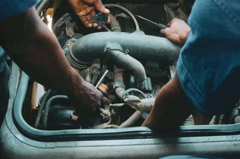 Mechanicy pracują przy silniku, jeden z nich trzyma wtyczkę, drugi dokręca śrubę. Może to być naprawa zaworu par paliwa.