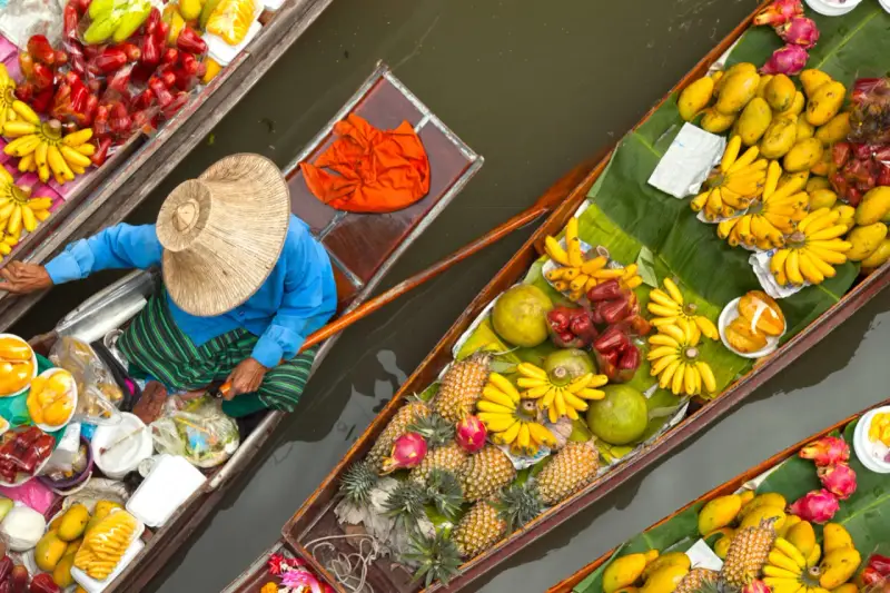 En säljare i en båt på en flytande marknad i Thailand, omgiven av ett överflöd av färsk frukt som bananer, mango och ananas.