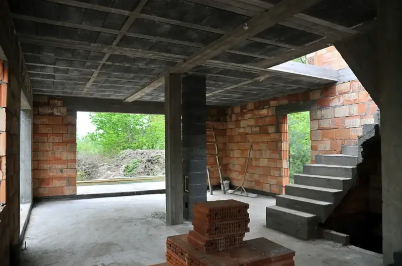 Wnętrze budowy z widocznym stropem gęstożebrowym, schodami, ceglanymi ścianami i otwartymi drzwiami na zewnątrz.