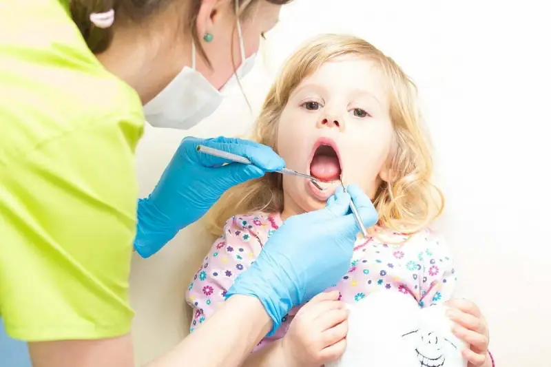 Mała dziewczynka z otwartymi ustami podczas pierwszej wizyty u dentysty. Dentystka w niebieskich rękawiczkach bada jej ząbki.