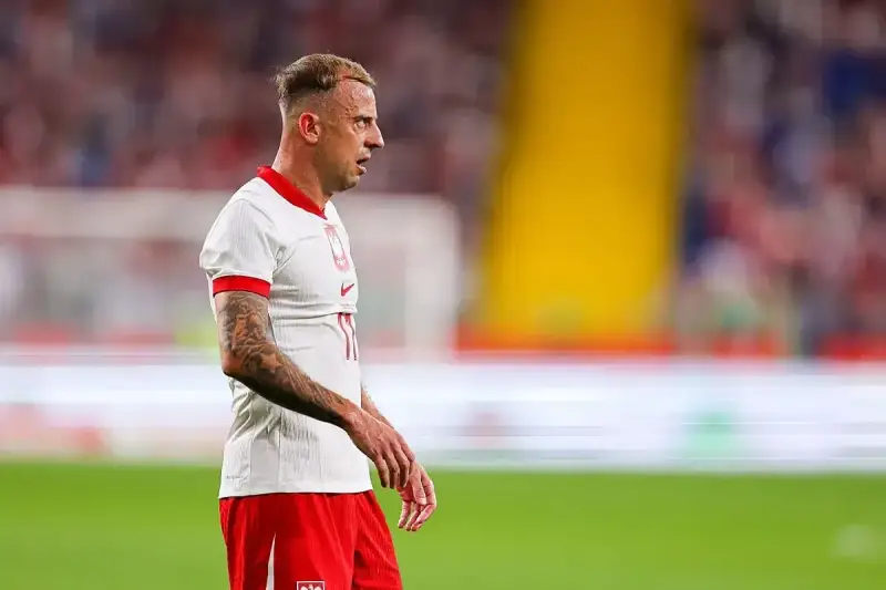 Kamil Grosicki w białej koszulce z orzełkiem i czerwonych spodenkach, gotowy do gry. Jego obecne drużyny to Polska i Pogoń Szczecin.