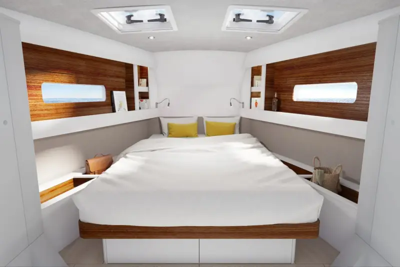 L'intérieur d'un voilier 8m, avec un grand lit blanc, des coussins jaunes, des hublots donnant sur la mer et des étagères en bois.