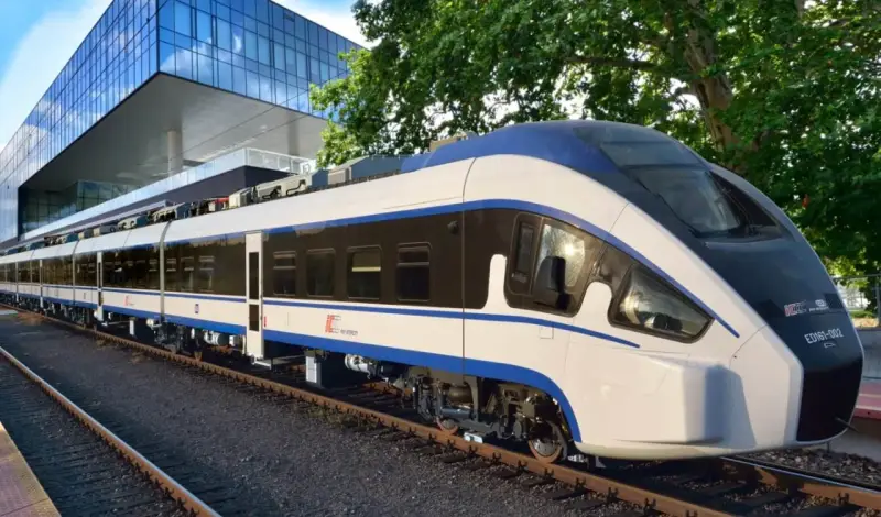 Nowoczesny, biało-niebieski pociąg Intercity ED161-002, najszybszy pociąg w Polsce, stoi na torach obok futurystycznego budynku.