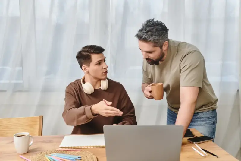 Ojciec rozmawia z synem, pokazując mu, jak zmotywować dziecko do nauki. Na stole laptop, zeszyt i kubek kawy.