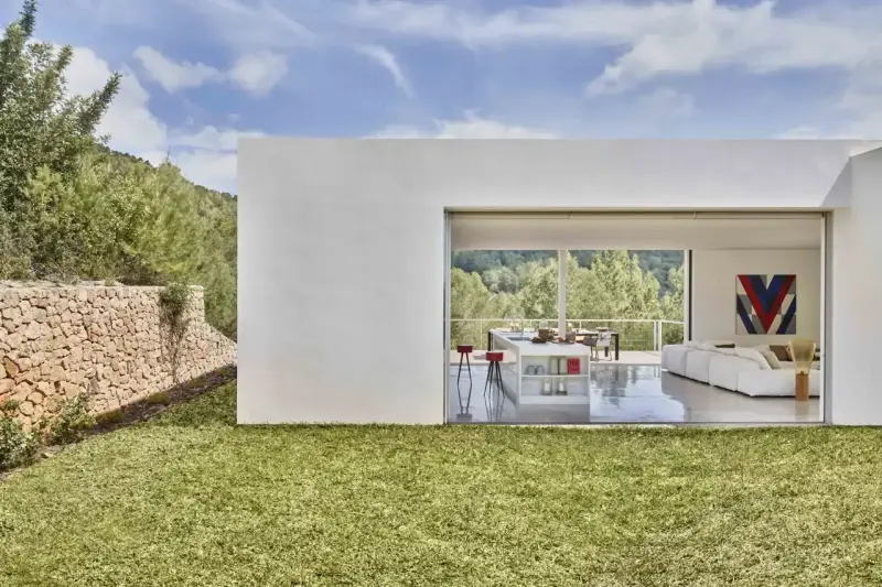 Exterior de casa ibicenca con césped verde y muro de piedra. Interior moderno con cocina y salón abiertos al paisaje.