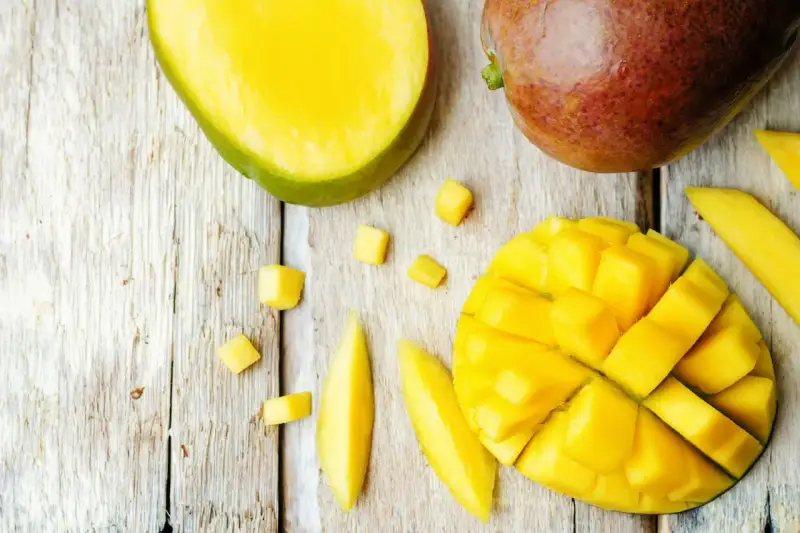 Czy pies może mango? Na drewnianym blacie leżą soczyste kawałki mango, pokrojone w kostkę i paski. Obok całe, dojrzałe owoce.