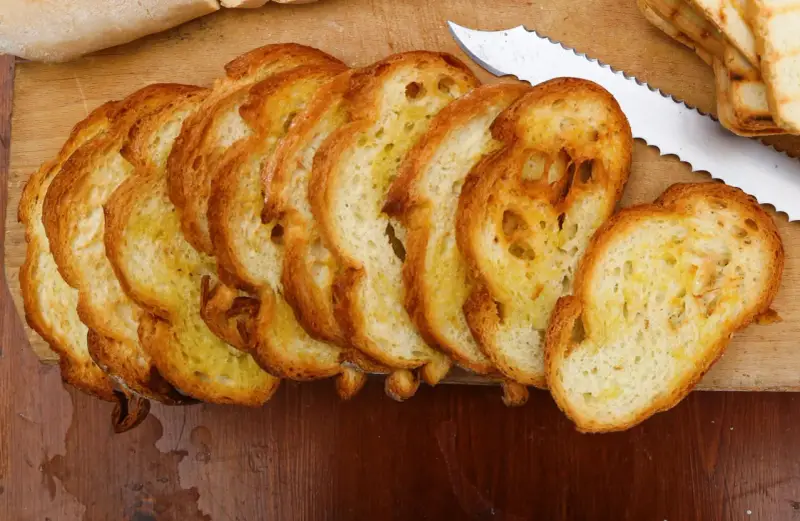 Fette dorate di pane con lievito madre, appena tostato, su un tagliere di legno. Un coltello seghettato è pronto per affettare altro pane.