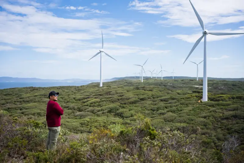 Mann steht vor Windrädern, die die australien energieversorgung repräsentieren. Die Landschaft ist grün und hügelig, mit dem Meer im Hintergrund.
