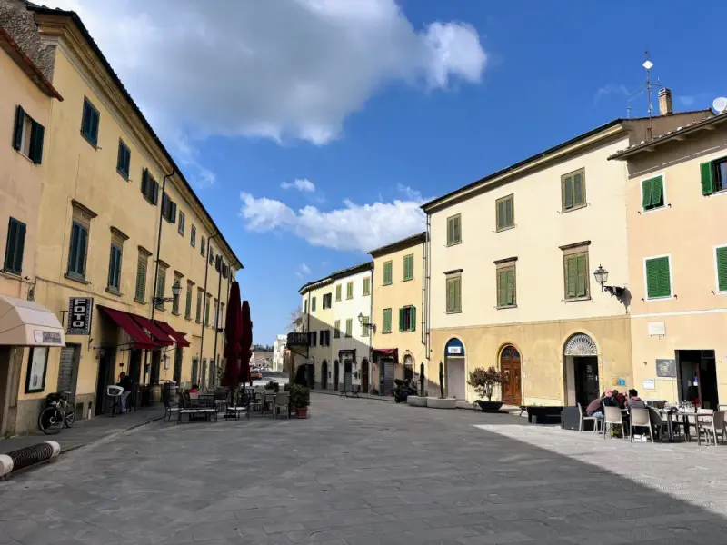 Piazza soleggiata a Pomarance, con edifici storici, tavolini all'aperto e scorci suggestivi da scoprire.