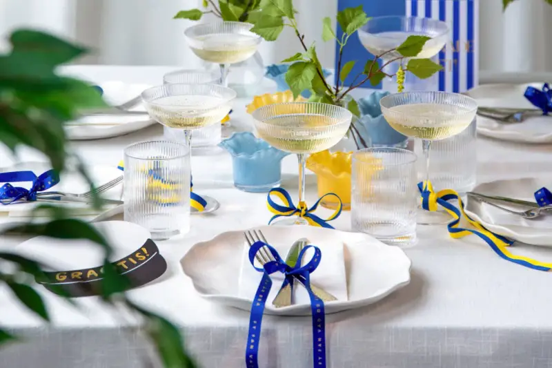 Festligt dukat bord med champagneglas, blå och gula band som knyts i en blå rosett, samt en studentmössa.