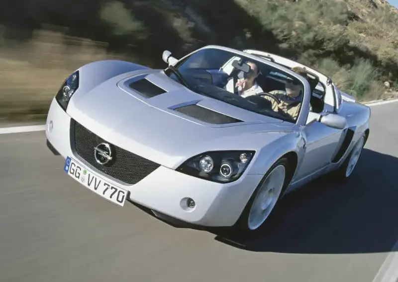 Srebrny Opel Speedster z dwoma pasażerami pędzi po krętej drodze.
