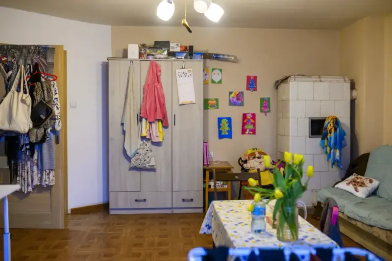 Przytulne mieszkanie dla samotnej matki. Szafa, piec kaflowy, kanapa, stolik z tulipanami i dziecięce rysunki na ścianie tworzą domową atmosferę.