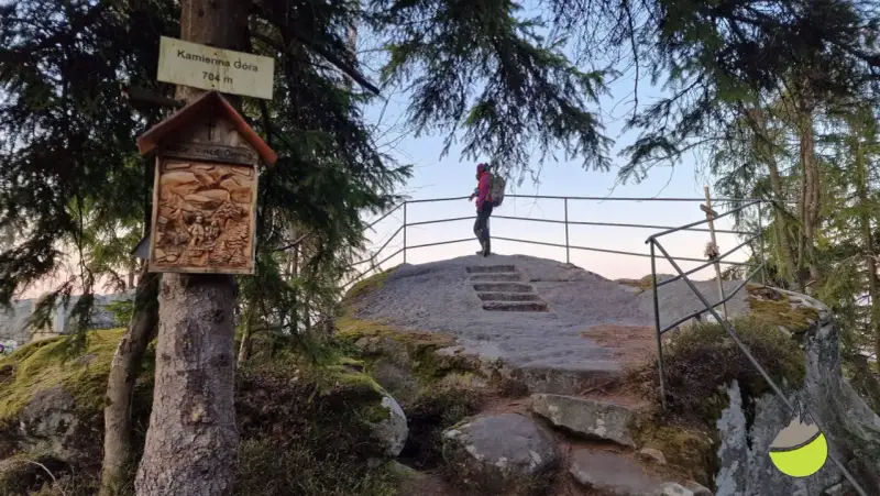 Widok na szczyt Kamienna Góra (704 m). Turystka podziwia panoramę, a obok drzewa ozdobiona kapliczka.