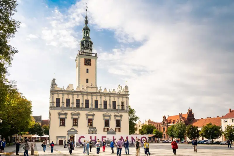 Rynek w Chełmnie z ratuszem i tablicą z napisem "Chełmno". Ludzie spacerują, podziwiając atrakcje miasta.