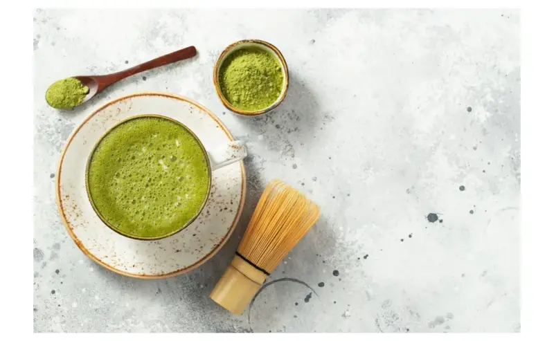 Zielona, spieniona matcha w filiżance, obok bambusowa miotełka i łyżeczka z proszkiem. To właśnie co to matcha – napój pełen energii.