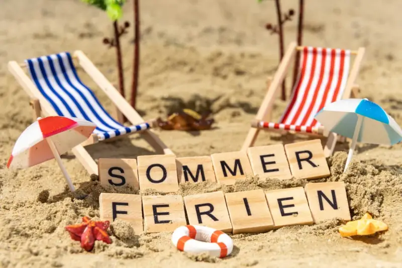 Strandkörbchen, Palmen und die Worte "Sommerferien" im Sand. Berlin wartet auf die Sommerferien 2026!