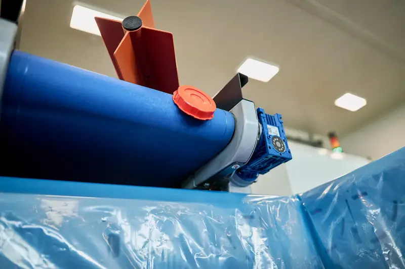 Maszyna do produkcji folii, z niebieskim wałkiem i pomarańczową nakrętką, przygotowuje materiał cerplast do dalszej obróbki.