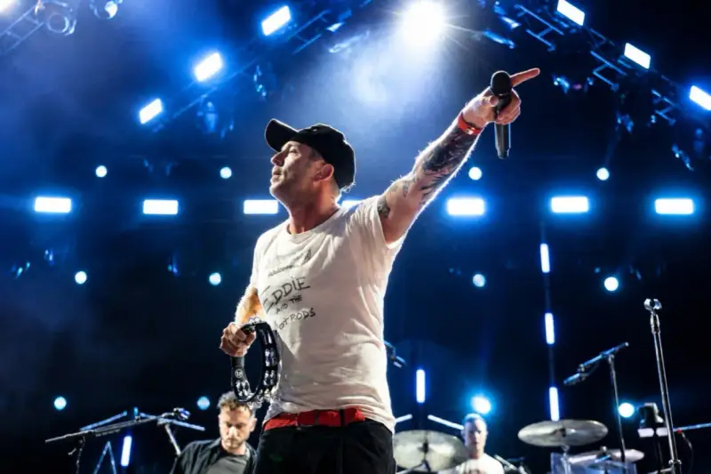 Wokalista z tatuażami i czapką na głowie prowadzi koncert One Republic, wskazując ręką w górę i trzymając tamburyn.