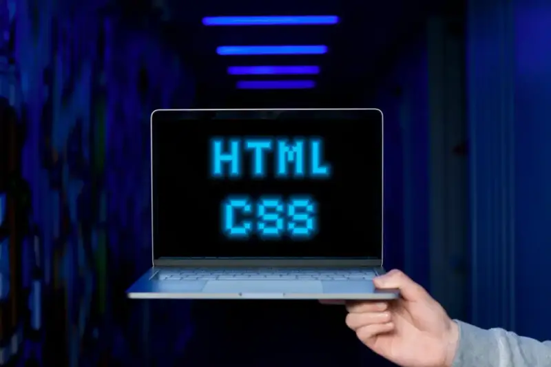 <p>Dłoń trzyma laptopa, na ekranie którego świeci napis "HTML CSS" w niebieskich pikselach. Tło jest ciemne, z niebieskimi neonami.</p>