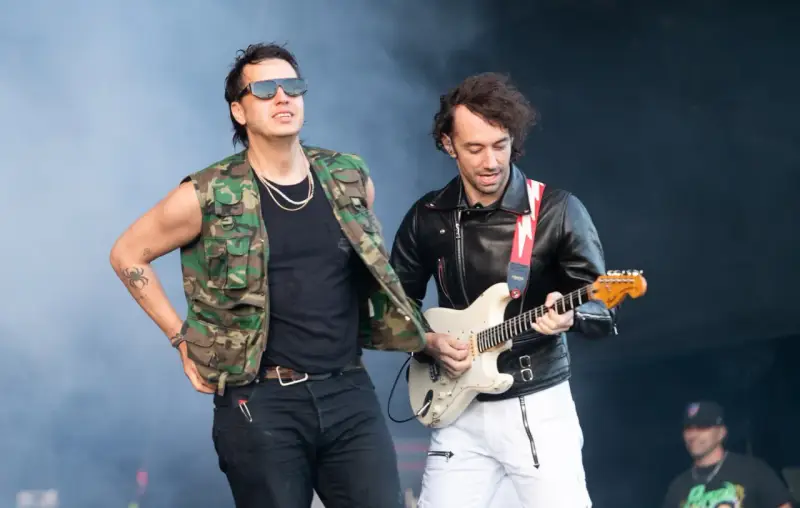 Albert Hammond Jr. w kamizelce moro i okularach stoi obok gitarzysty w skórzanej kurtce, grającego na białej gitarze.