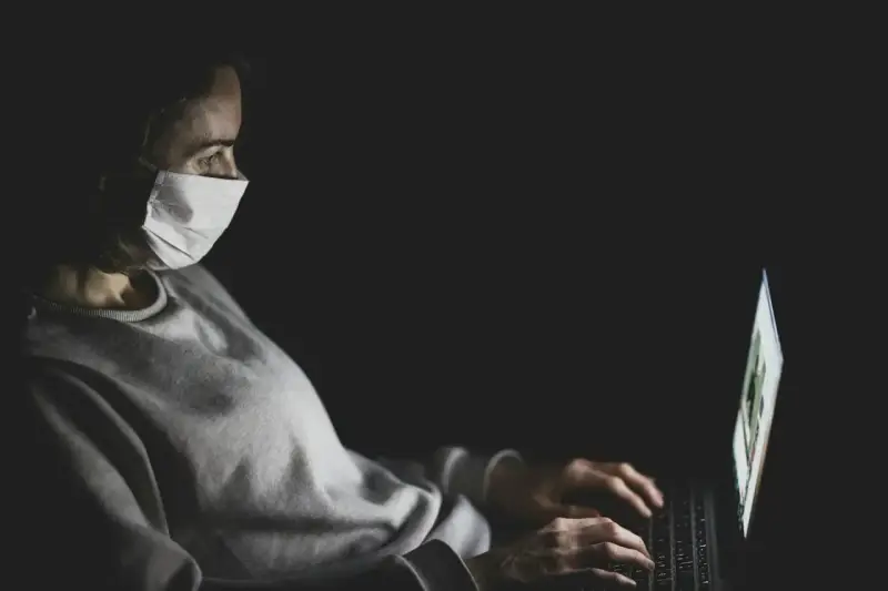 Kobieta w masce medycznej pracuje na laptopie w ciemności.