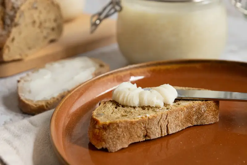 Ein Glas mit selbstgemachtem Schmalz steht bereit. Eine Scheibe Brot wird gerade mit dem Schmalz bestrichen, ein Genuss für jeden, der Schmalz selber machen möchte.
