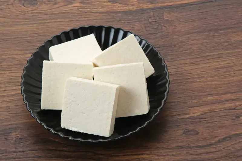 Kilka kostek białego tofu na czarnym talerzu. Czy tofu można mrozić? Tak, po rozmrożeniu zmienia teksturę, stając się bardziej gąbczaste.