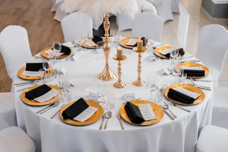 Elegancko nakryty stół weselny ze złotymi talerzami, czarnymi serwetkami i świecami. Wesele motywy złota i bieli.