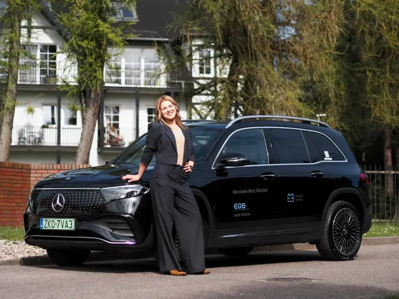 Kobieta w czarnym garniturze opiera się o czarnego Mercedesa EQB na tle białego domu.