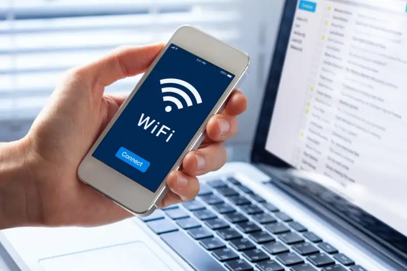 Dłoń trzyma smartfon z ikoną WiFi i napisem "Connect". Obok widać laptopa z listą e-maili, sugerującą pracę zdalną i dostęp do sieci, być może przez **ssid** domowy.
