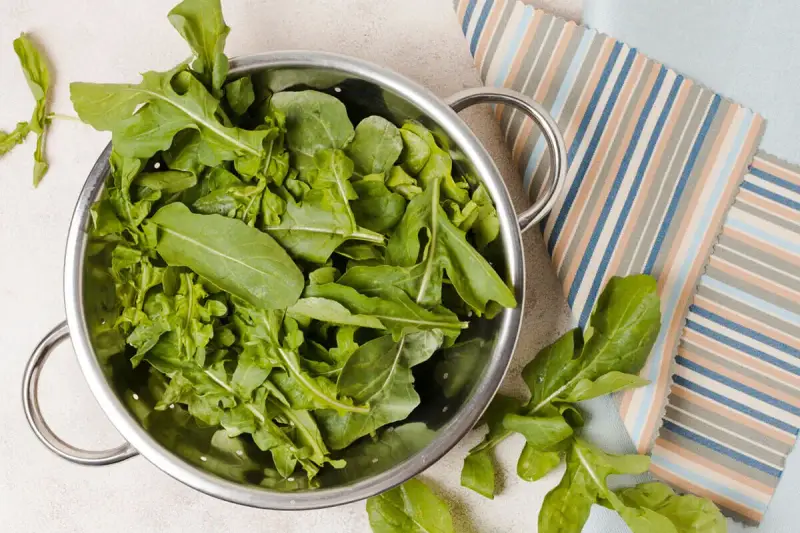 Rukola w durszlaku, idealna do przygotowania jadłospisu tygodniowego dieta FODMAP. Świeże liście na tle kolorowych tkanin.