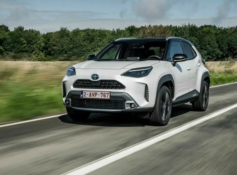 Biały Yaris Cross Hybrid jedzie po drodze, otoczony zielenią.