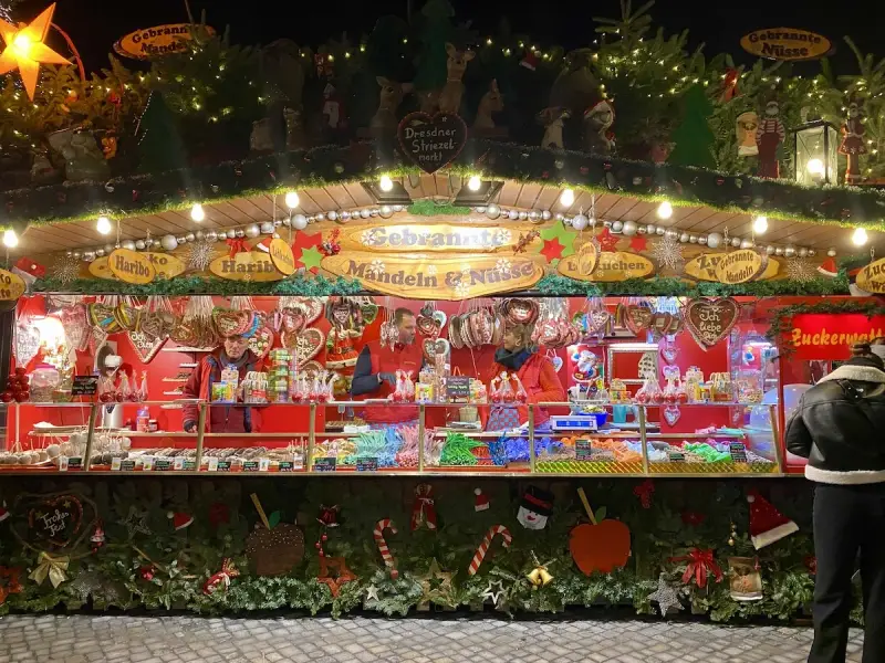 A vibrant Dresden Christmas market stall overflowing with gingerbread hearts, candied nuts, and colorful sweets. Festive lights twinkle above the tempting displays.