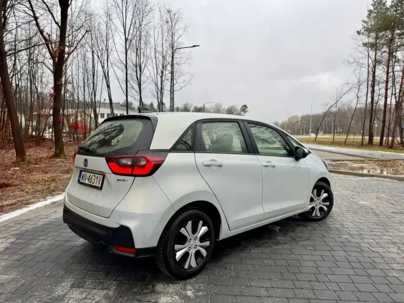Biały Honda Fit stoi na mokrej, brukowanej nawierzchni. W tle drzewa i budynki.